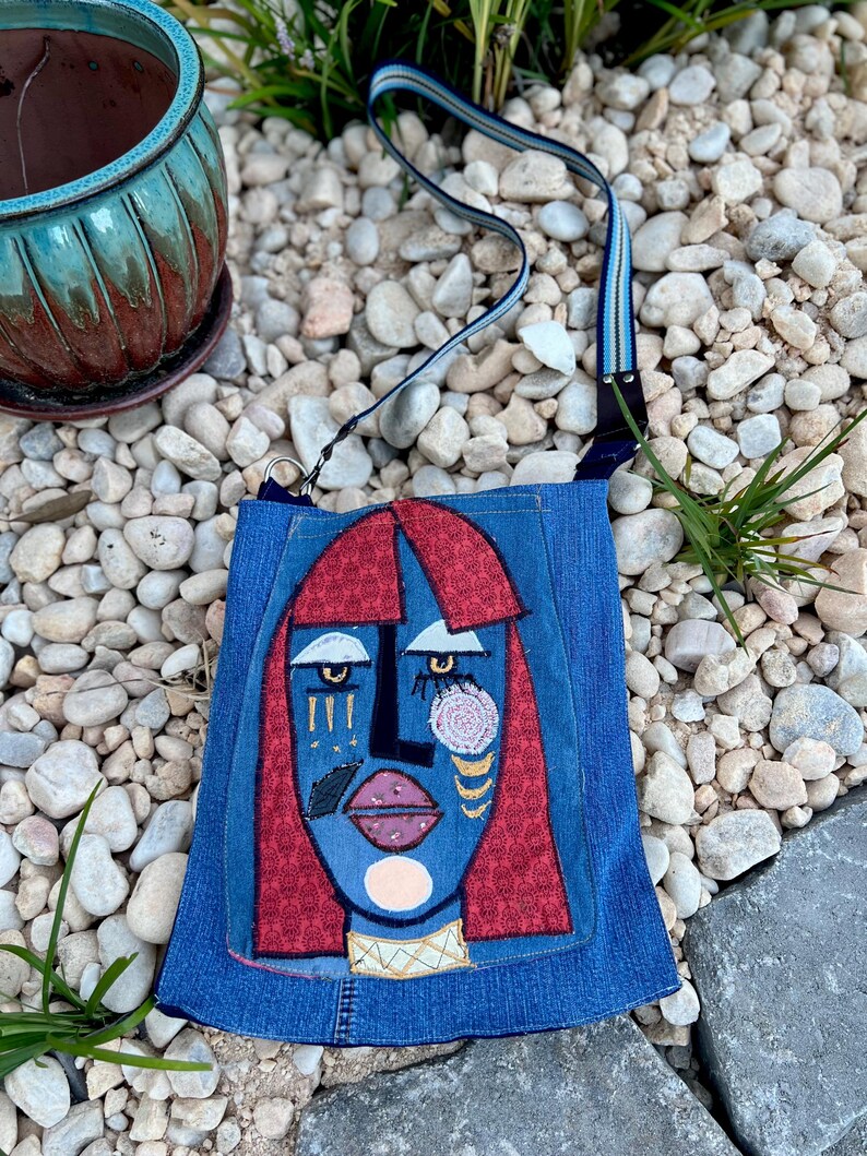 May include: A denim crossbody bag with an abstract face design. The face has red hair, blue skin, and geometric shapes. The bag has a striped strap. The background features pebbles and greenery.