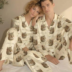 May include: Cream-colored pajama set featuring a repeating pattern of cat faces and the text "Bella's Meow." The set includes a button-down top and pants. The pajamas are made of a soft, silky material.