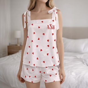 Puede incluir: Conjunto de pijama blanco con estampado de corazones rojos. La parte superior tiene tirantes anudados y las iniciales "ASH" en rojo. Los pantalones cortos completan el conjunto. Esta ropa de dormir es ideal para una noche acogedora.