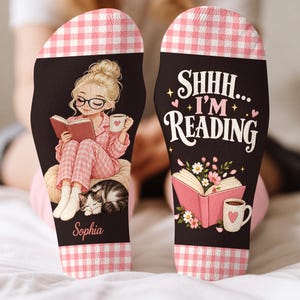 Puede incluir: Un par de calcetines negros con puños de cuadros rosas y blancos. Un calcetín muestra una ilustración de una persona leyendo con un gato y el nombre Sophia. El otro muestra el texto SHHH... I'M READING con un libro abierto y una taza.