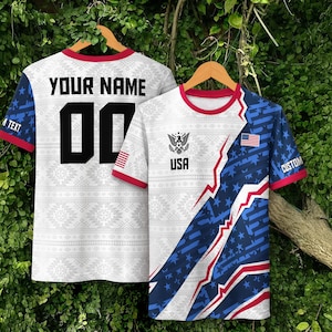 May include: Two short-sleeve t-shirts with red trim. The left shirt features 'YOUR NAME' and '00' in black. The right shirt displays a white, blue, and red pattern with stars, a USA flag, an eagle emblem, and the text 'USA' and 'CUSTOM'.