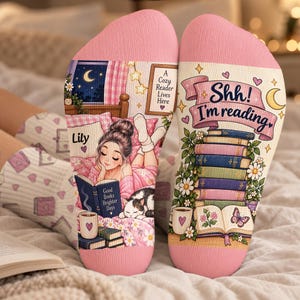 May include: A pair of pink-topped socks featuring a printed illustration of a person reading in bed with a cat, the text 'Lily', 'A Cozy Reader Lives Here', 'Good Books Brighter Days', and 'Shh! I'm reading' above a stack of colourful books.