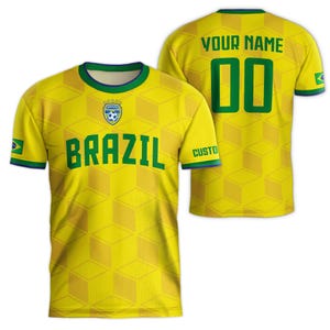 May include: Yellow soccer jersey with green and blue accents. The front displays the word "BRAZIL" in green block letters, a soccer ball emblem, and the Brazilian flag. The back features the text "YOUR NAME" and the number "00" in green.