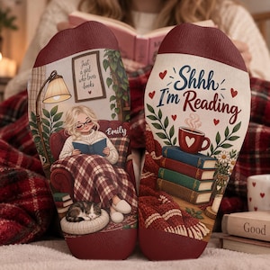 May include: A pair of socks featuring a maroon cuff and a graphic print. One sock shows a person reading in a chair with a cat, a lamp, and a sign reading 'Just a girl who loves books'. The other sock displays the text 'Shhh I'm Reading' with a stack of books, a steaming mug, and small red hearts.