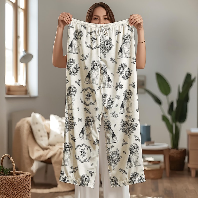 K&ouml;nnte beinhalten: Cremefarbene Schlafhose mit einem sich wiederholenden Muster aus Beagle-Illustrationen und floralen Akzenten in Schwarz und Wei&szlig;. Die Hose hat einen Kordelzug in der Taille und eine lockere Passform. Der Stoff wirkt weich und leicht.