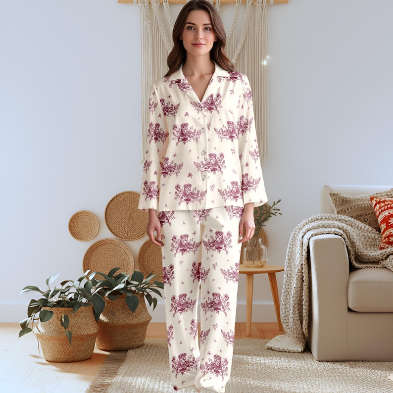 May include: A two-piece pajama set featuring a long-sleeved button-down shirt and matching loose-fitting trousers. The fabric is cream-colored with a repeating maroon-toned toile pattern depicting various animals and floral elements throughout.