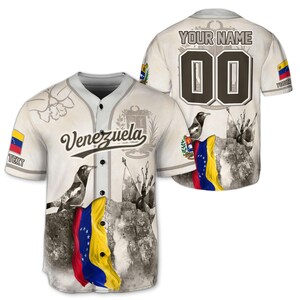 May include: A baseball jersey with the word "Venezuela" in script. The jersey is off-white with a gray collar and buttons. The front features a flag graphic and a bird. The back has the text "YOUR NAME" above the number "00".