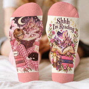 May include: A pair of pink socks featuring a printed illustration of a person reading a book with a cat, a castle, and the text 'Shhh I'm Reading'. The design includes stacks of books, a candle, flowers, and a crescent moon on a light background.