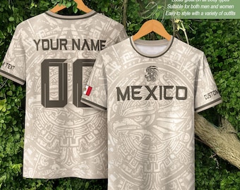 May include: Two beige t-shirts with a repeating Aztec-inspired pattern. The front features the word MEXICO, an eagle graphic, and a small Mexican flag. The back displays the text YOUR NAME and the number 00. Both shirts are on wooden hangers.