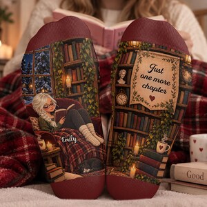 May include: A pair of maroon socks featuring a printed scene of a person reading in a cosy library with a cat, a window showing a snowy night, and the name Emily. The other sock displays a bookshelf with the text Just one more chapter and a heart.