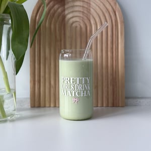 Op de afbeelding: Een glas met een groene drank en een glazen rietje. Het glas heeft de tekst "PRETTY GIRLS DRINK MATCHA" erop gedrukt met een roze strik.