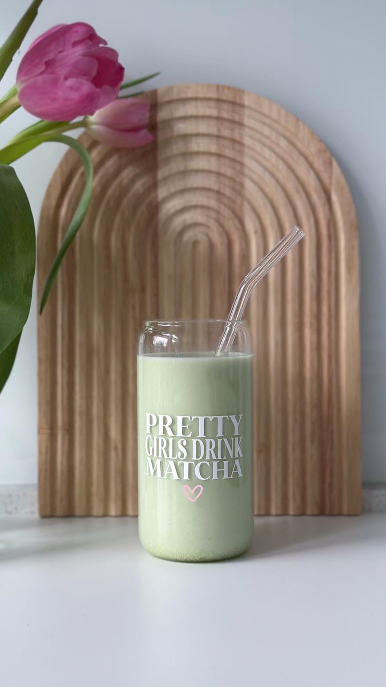 Op de afbeelding: Een glas met een groene drank en een doorzichtige rietje. Het glas heeft de tekst "PRETTY GIRLS DRINK MATCHA" met een roze hart.