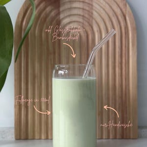 May include: A clear glass with a green liquid inside, a glass straw, and a bamboo lid. The glass is sitting on a wooden surface. The text "inkl. Glasstrohhalm + Bambusdeckel" is written above the glass. The text "F&uuml;llmenge ca. 500ml" is written below the glass. The text "nur Handw&auml;sche" is written to the right of the glass.