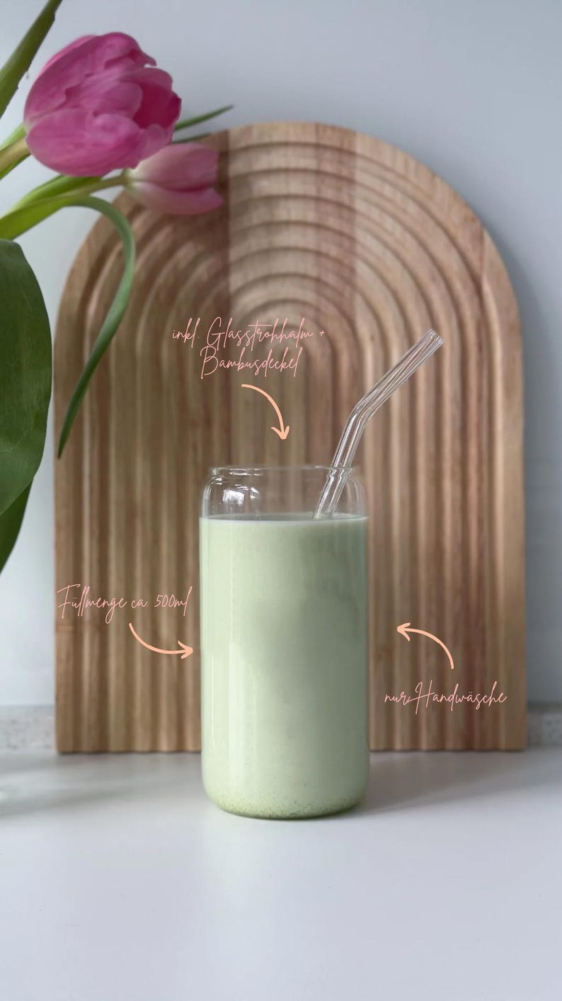 May include: A clear glass with a green liquid inside, a glass straw, and a bamboo lid. The glass is sitting on a wooden surface. The text "inkl. Glasstrohhalm + Bambusdeckel" is written above the glass. The text "F&uuml;llmenge ca. 500ml" is written below the glass. The text "nur Handw&auml;sche" is written to the right of the glass.