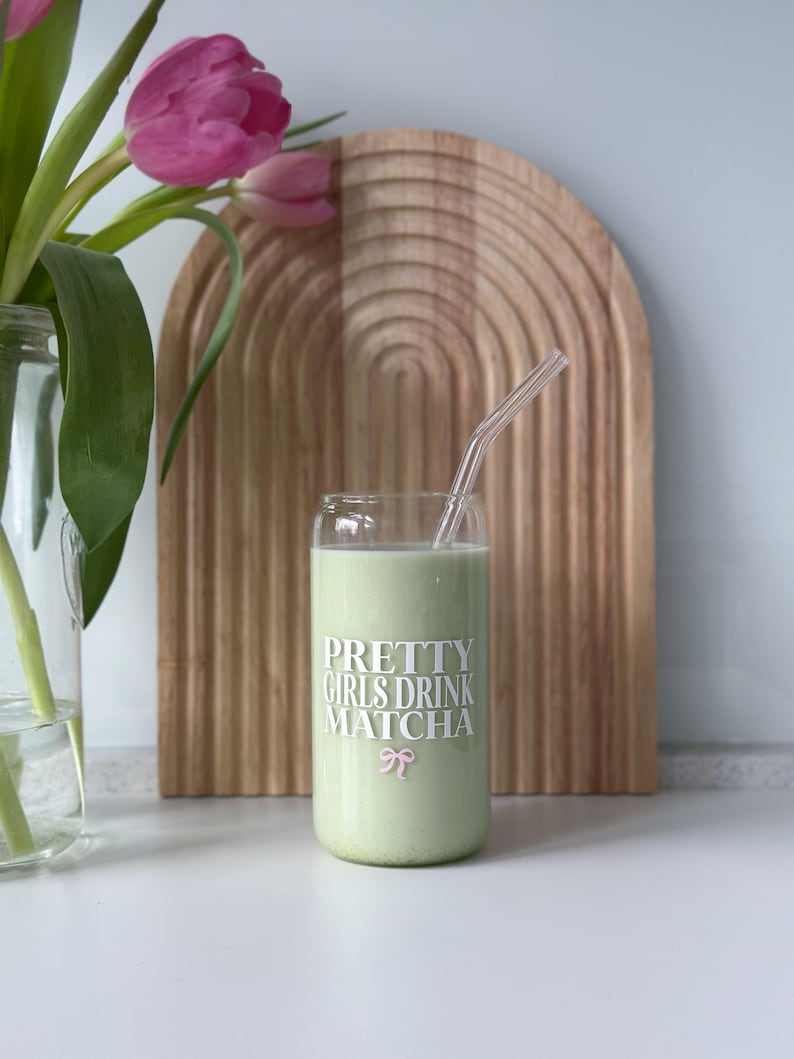 Op de afbeelding: Een glas met een groene drank en een glazen rietje. Het glas heeft de tekst "PRETTY GIRLS DRINK MATCHA" erop gedrukt met een roze strik.