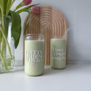 Op de afbeelding: Twee groene glazen bekers met rietjes, gevuld met een groene drank. De bekers hebben witte tekst die "Pretty Girls Drink Matcha" en "I love you, so matcha" luidt.