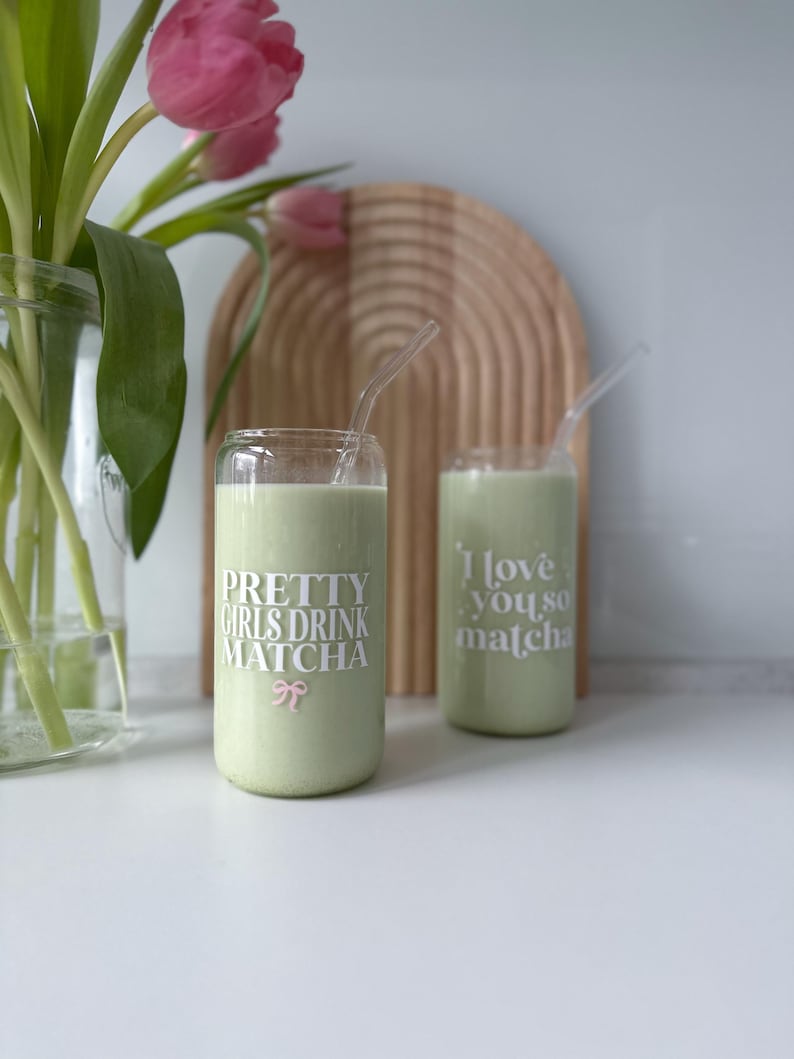 Op de afbeelding: Twee groene glazen bekers met rietjes, gevuld met een groene drank. De bekers hebben witte tekst die "Pretty Girls Drink Matcha" en "I love you, so matcha" luidt.