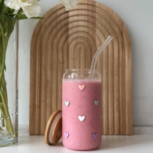 May include: A glass jar filled with a pink smoothie and a glass straw. The jar has a wooden lid and is decorated with colourful hearts.