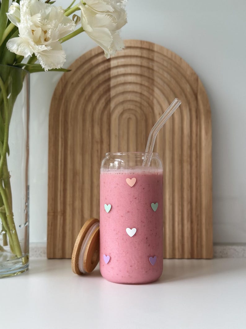 May include: A glass jar filled with a pink smoothie and a glass straw. The jar has a wooden lid and is decorated with colourful hearts.