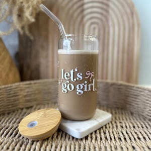 Könnte beinhalten: Ein Glasbecher mit Bambusdeckel und Glasstrohhalm. Der Becher enthält eine braune Flüssigkeit und den Text "let's go girl" auf der Seite.