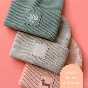 Könnte beinhalten: Drei gerippte Strickmützen in Salbeigrün, Beige und Altrosa. Jede Mütze hat einen quadratischen Stoffaufnäher am Umschlag. Die grüne Mütze zeigt den Text "BAD HAIR day", die beige "mausig" und die rosa Mütze eine Grafik eines Dackels.