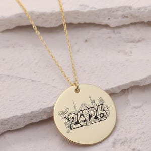 May include: A gold-colored necklace with a circular pendant. The pendant features a black and white illustration of landmarks and the number "2026". The necklace chain is delicate and gold-toned, with the pendant hanging in the center.