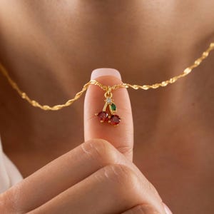 Könnte beinhalten: Eine goldfarbene Halskette mit einem Kirschenanhänger. Der Anhänger hat zwei rote Edelsteine für die Kirschen, einen grünen Edelstein für das Blatt und einen kleinen klaren Edelstein. Die Kette hat ein gedrehtes Seildesign.
