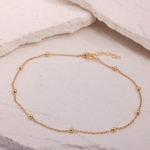Op de afbeelding: Een delicate goudkleurige enkelband met kleine, gelijkmatig verdeelde bolvormige kralen. De enkelband is gemaakt van een fijne ketting en heeft een verstelbare sluiting. De sieraden worden gepresenteerd op een neutrale, gebroken witte ondergrond.