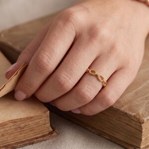 Könnte beinhalten: Ein goldener Ring, bestehend aus einem durchgehenden Band aus kleinen, ovalen Gliedern mit einer texturierten, perlenartigen Oberfläche. Der Ring wird an einem Finger getragen und zeigt die filigrane Metallarbeit des ovalen Designs.