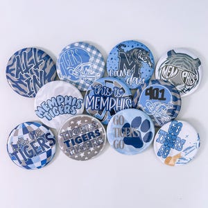 May include: A collection of round buttons with various designs in shades of blue and white. Some buttons feature text like "ALL IN," "TIGERS," and "MEMPHIS." Other designs include a tiger, a football helmet, and a paw print.