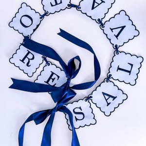 Może przedstawiać: Niebiesko-biała girlanda z literami "OREVALISA" w falistym wzorze. Girlanda jest związana niebieską wstążką.