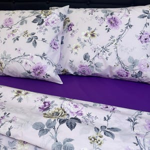 May include: Bed linen set with a light pink floral design, showcasing purple and white roses and grey leaves. The set includes two pillowcases and a sheet, set against a deep purple sheet and a black headboard.