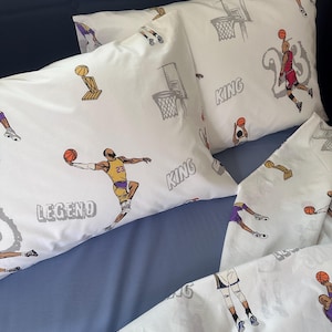May include: White basketball-themed bedding set with illustrations of basketball players, hoops, and trophies. The pillowcases have the words "KING" and "LEGEND". The set includes a fitted sheet and pillowcases.