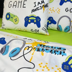 Könnte beinhalten: Weißes Bettwäsche-Set mit Gaming-Motiv. Das Set enthält zwei Kissen und ein Laken, verziert mit Gamecontrollern, Kopfhörern und den Schriftzügen „GAME“, „I AM AWESOME“ und „MAKE SOME NOISE“. Das Design weist blaue, grüne und gelbe Akzente auf.