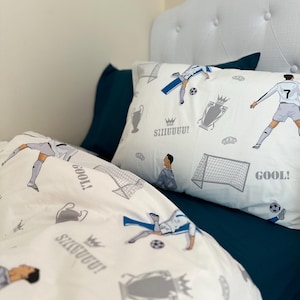 May include: Football-themed bedding set in white, featuring a duvet cover and pillowcase. The design includes illustrations of a soccer player, goals, trophies, and the word "GOOL!". The bedding is set on a dark blue sheet.