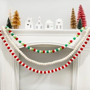 Peut inclure: Un manteau blanc d&eacute;cor&eacute; de trois guirlandes de boules en feutre rouges, vertes et blanches. De petites maisons blanches et des sapins de No&euml;l de diff&eacute;rentes couleurs sont dispos&eacute;s sur le manteau, cr&eacute;ant une sc&egrave;ne de f&ecirc;te.