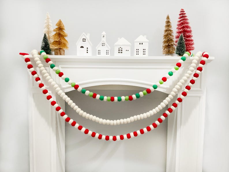 Peut inclure: Un manteau blanc d&eacute;cor&eacute; de trois guirlandes de boules en feutre rouges, vertes et blanches. De petites maisons blanches et des sapins de No&euml;l de diff&eacute;rentes couleurs sont dispos&eacute;s sur le manteau, cr&eacute;ant une sc&egrave;ne de f&ecirc;te.