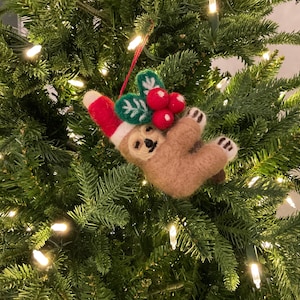 May include: A handmade Christmas tree ornament featuring a brown sloth wearing a red and white Santa hat. The sloth is holding green holly leaves and red berries. The ornament hangs on a green Christmas tree with white lights.
