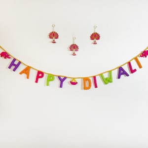 May include: A festive Diwali decoration featuring a garland spelling "HAPPY DIWALI" in colorful letters. The garland is adorned with pink elephants and hangs below lotus flower ornaments and white beaded strands.