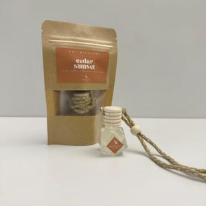 May include: A brown paper bag with a white label that reads "cedar sunset" and a small glass bottle with a wooden cap and a gold cord hanging from it. The bottle is filled with a clear liquid and has a label that reads "cedar sunset".