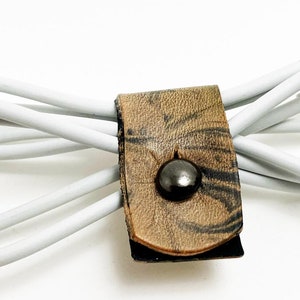 May include: A brown leather cord organizer with a black button. A white charging cable is wrapped around the organizer.