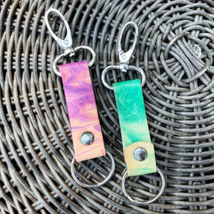 May include: Two leather keyrings with silver key rings. One keyring is pink and purple with a swirl pattern. The other keyring is green and yellow with a swirl pattern.