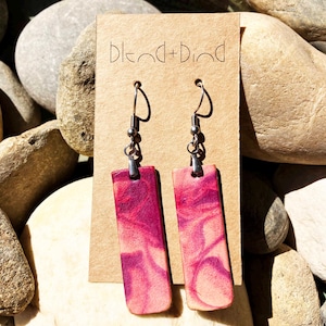 Puede incluir: Un par de pendientes de madera rosa y blanca con ganchos plateados. Los pendientes son de forma rectangular y tienen un patrón de mármol. Los pendientes se muestran en una tarjeta marrón con el texto "Blend + Bind".