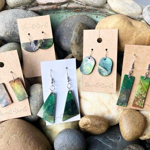 May include: Four pairs of dangle earrings with green and brown abstract designs on a background of brown rocks. The earrings are labeled "blend+bind".