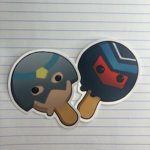 Può includere: Due adesivi con disegni di ghiaccioli a fumetti. Uno ha un design a casco blu e grigio con un accento giallo, l'altro ha un ghiacciolo blu scuro con un design a maschera rossa. Entrambi hanno bastoncini marroni.