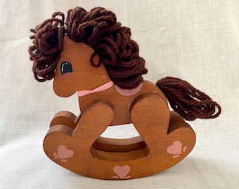May include: A wooden rocking horse with a brown finish, featuring a dark brown yarn mane and tail. The horse has a pink collar and pink heart accents on the rockers. The toy is designed for young children.