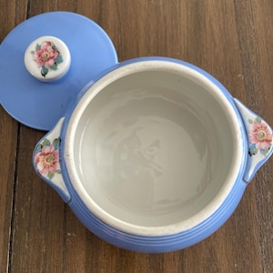 Può includere: Una ciotola da portata in ceramica azzurra con coperchio abbinato. Il coperchio presenta un motivo floreale sul pomello. La ciotola ha due manici e decorazioni floreali. L'interno è bianco.