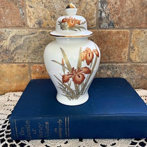 May include: White porcelain ginger jar with a lid and a floral design of orange and gold irises. The jar is sitting on a blue book titled "The Education of Henry Adams".