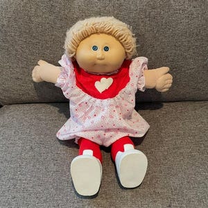 May include: A vintage doll with a soft, round face, blue eyes, and blonde yarn hair. The doll wears a red and white dress with heart patterns, red leggings, and white shoes. The doll's arms are outstretched.