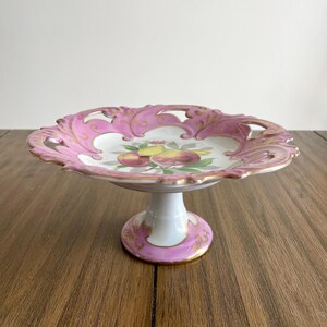 Pode incluir: Uma travessa vintage com pedestal, com base branca e borda rosa ondulada. A borda apresenta uma moldura dourada e um design vazado ornamentado. O centro da travessa exibe um motivo de frutas pintadas.